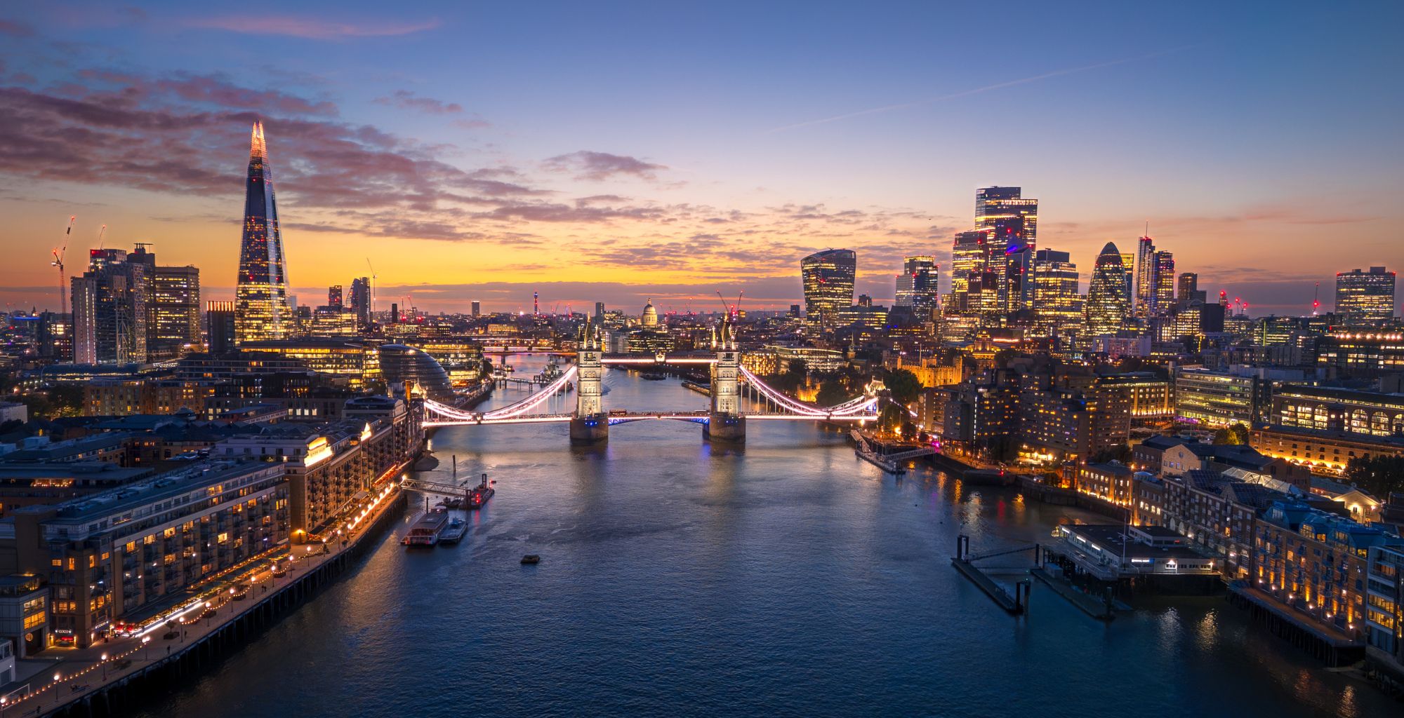 Evening cityscape of London with Tower Bridge lit up over the Thames at sunset, skyscrapers in the background.